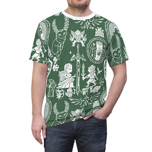 Op de afbeelding: Een groen T-shirt met een witte print van verschillende symbolen en karakters, waaronder een zwaard, een hart, een vogel en een hert. De print is een herhalend patroon.