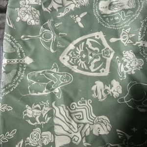 May include: A sage green fabric featuring a repeating white graphic print of various fantasy-themed icons, including a shield with a central triangular emblem, a sword, circular runic patterns, and stylized figures of characters in motion.