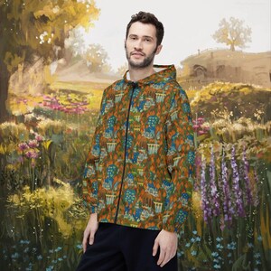 May include: A zip-up hooded jacket with a colorful pattern of houses and trees in shades of orange, green, and blue. The jacket has a black zipper and a white interior. The background is a painting of a landscape with flowers and trees.