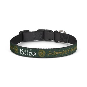 May include: A dark green pet collar featuring the white text Bilbo, gold circular star motifs, and gold elvish script. The collar includes a black plastic side-release buckle and a silver-toned metal D-ring for attaching a leash or identification tag.
