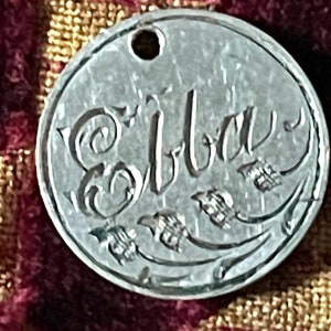 May include: A silver charm with the name "Elba" engraved in a cursive script. The charm has a hole at the top for attaching to a necklace or bracelet. The background is a blurry image of a red and gold fabric.