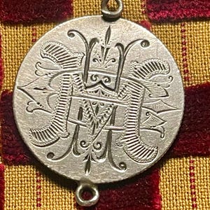 May include: A circular silver-toned metal pendant featuring an intricate engraved monogram of the letters M and H intertwined. The disc has a small circular loop at both the top and the bottom for attachment, with detailed decorative line work.