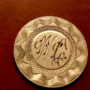 May include: A silver-colored token with a wavy border and a central design featuring a monogram of the letters "W" and "H" in cursive script.