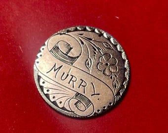 May include: A silver-colored button with the word "MURRY" engraved on a banner. The button features floral and scrollwork designs around the edge and on the banner.