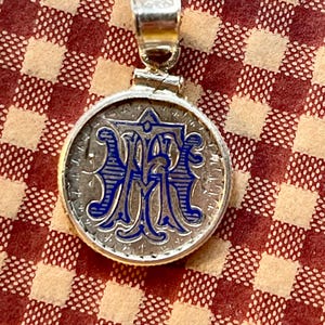May include: A silver pendant with a blue and silver monogram. The pendant is round and has a bail at the top.