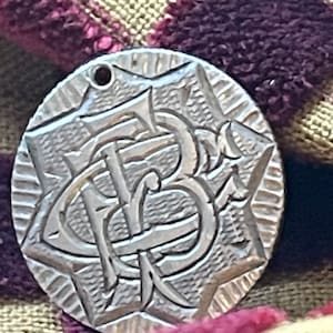 May include: A silver oval-shaped pendant with a detailed embossed design of the letters "FB" intertwined. The pendant has a small hole at the top for attaching a chain.