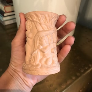 May include: A terracotta vase with a detailed design of cherubs and a tree. The vase is a light brown color and has a smooth, matte finish.