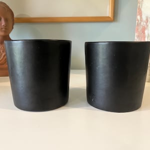 May include: Two black ceramic cups on a white surface. The cups are cylindrical and have a smooth, matte finish.