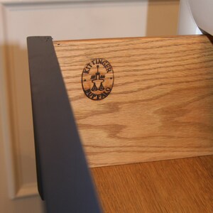 May include: Close-up of a wooden drawer with a black Kittinger Buffalo logo. The drawer is made of light wood with a grain pattern.