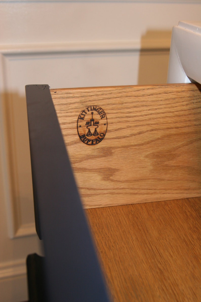 May include: Close-up of a wooden drawer with a black Kittinger Buffalo logo. The drawer is made of light wood with a grain pattern.