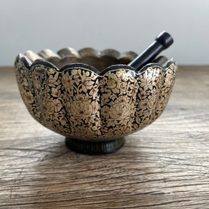 May include: A black and gold painted wooden bowl with a floral design and a matching wooden pestle. The bowl has a scalloped edge and sits on a wooden surface.