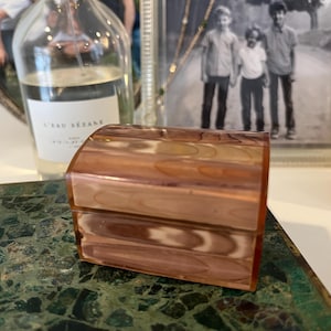 May include: A small, wooden box with a hinged lid. The box is made of light brown wood with a marbled pattern. The box is sitting on a green and brown surface.