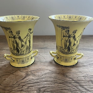 May include: Two yellow ceramic vases with black and white designs of women in a garden setting. The vases have two handles on each side.