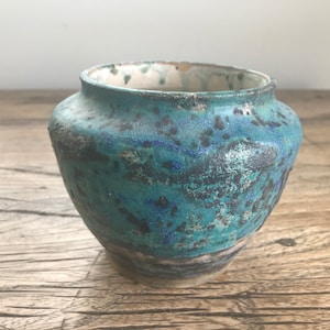 May include: A small, teal blue ceramic vase with a speckled glaze. The vase has a rounded body and a slightly flared rim.
