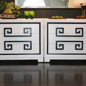 May include: Two white dressers with black trim and Greek key design. Each dresser has three drawers.