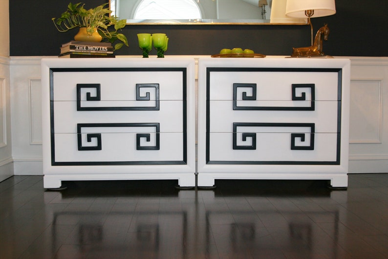 May include: Two white dressers with black trim and Greek key design. Each dresser has three drawers.