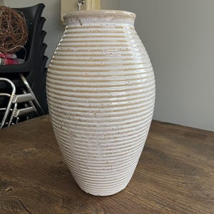 May include: A cream-colored ceramic vase with horizontal ridged detailing. The vase has a wide body that tapers towards the top and bottom. The rim of the vase is a lighter shade of cream. The vase is sitting on a wooden surface.