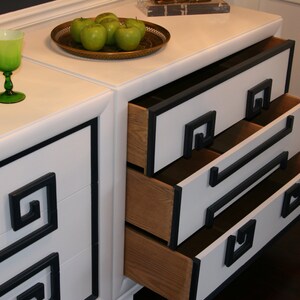 May include: White dresser with three drawers and black trim. The drawers have a geometric design on the front and are open, revealing the wooden interior. Green apples are on the top of the dresser.