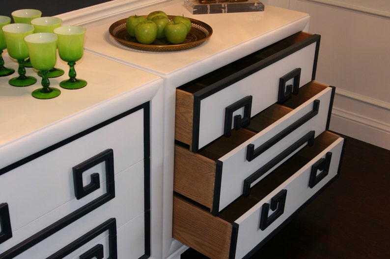 May include: White dresser with three drawers and black trim. The drawers have a geometric design on the front and are open, revealing the wooden interior. Green apples are on the top of the dresser.