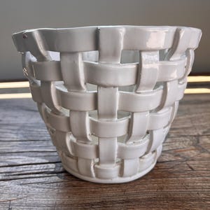 May include: A white ceramic planter featuring a woven basket-style design with an open lattice pattern. The structure consists of interconnected, smooth, glossy strips of ceramic that form a cylindrical shape with a solid base and an open top.