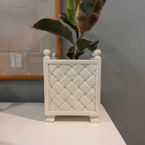 May include: A square white ceramic planter featuring a raised basket-weave texture on its sides. The planter has four small feet at the base and decorative spherical finials at each of the four top corners, containing a variegated rubber plant.