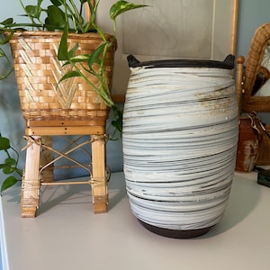 May include: A woven bamboo planter with a green plant and a large ceramic vase. The vase is white with horizontal gray and brown stripes and has two small handles. The planter is on a bamboo stand.