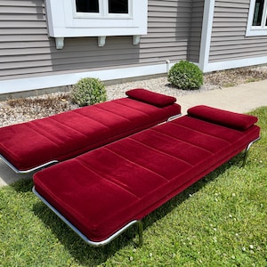 May include: Two red velvet chaise lounges with chrome frames are positioned on a grassy lawn. Each lounge has a rectangular seat and a padded headrest. The lounges are outdoors, with a house and landscaping in the background.