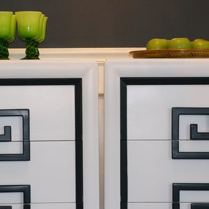 May include: Two white dressers with black trim and a Greek key design. The dressers have three drawers each. The top of the dressers is white and has a black trim. There are green glasses on the top of the dressers.