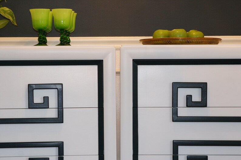 May include: Two white dressers with black trim and a Greek key design. The dressers have three drawers each. The top of the dressers is white and has a black trim. There are green glasses on the top of the dressers.