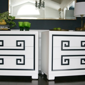 May include: Two white dressers with black trim and Greek key design. Each dresser has three drawers. The dressers are sitting on a dark wood floor.