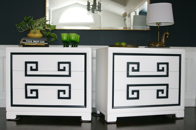 May include: Two white dressers with black trim and Greek key design. Each dresser has three drawers. The dressers are sitting on a dark wood floor.