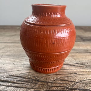 May include: A small, terracotta-colored ceramic vase with a textured, patterned surface. The vase has a rounded body and a slightly flared lip.