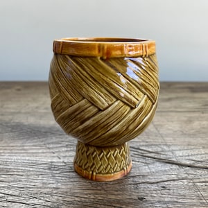 May include: A brown ceramic tiki mug shaped like a woven basket with a brown and orange rim. The mug has a woven pattern and stands on a small base.