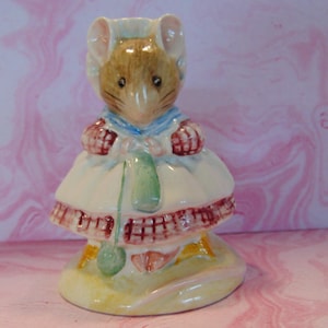 May include: A ceramic figurine of a mouse wearing a bonnet and dress. The mouse has brown fur, pink ears, and a blue collar. The dress is white with a red and brown checkered trim. The figurine is on a yellow base.