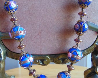 Könnte beinhalten: Eine Vintage-Halskette mit großen, runden blauen Glasperlen, kupferfarbenen Akzenten und rosa und weißen floralen Designs. Die Perlen sind mit kleineren kupferfarbenen Perlen und Verbindern aufgefädelt, was ein einzigartiges Schmuckstück ergibt.