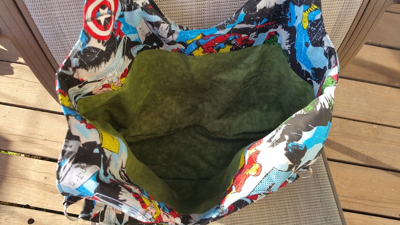 May include: A large green fabric tote bag with a superhero comic book print. The bag is open and the inside is lined with a dark green fabric.