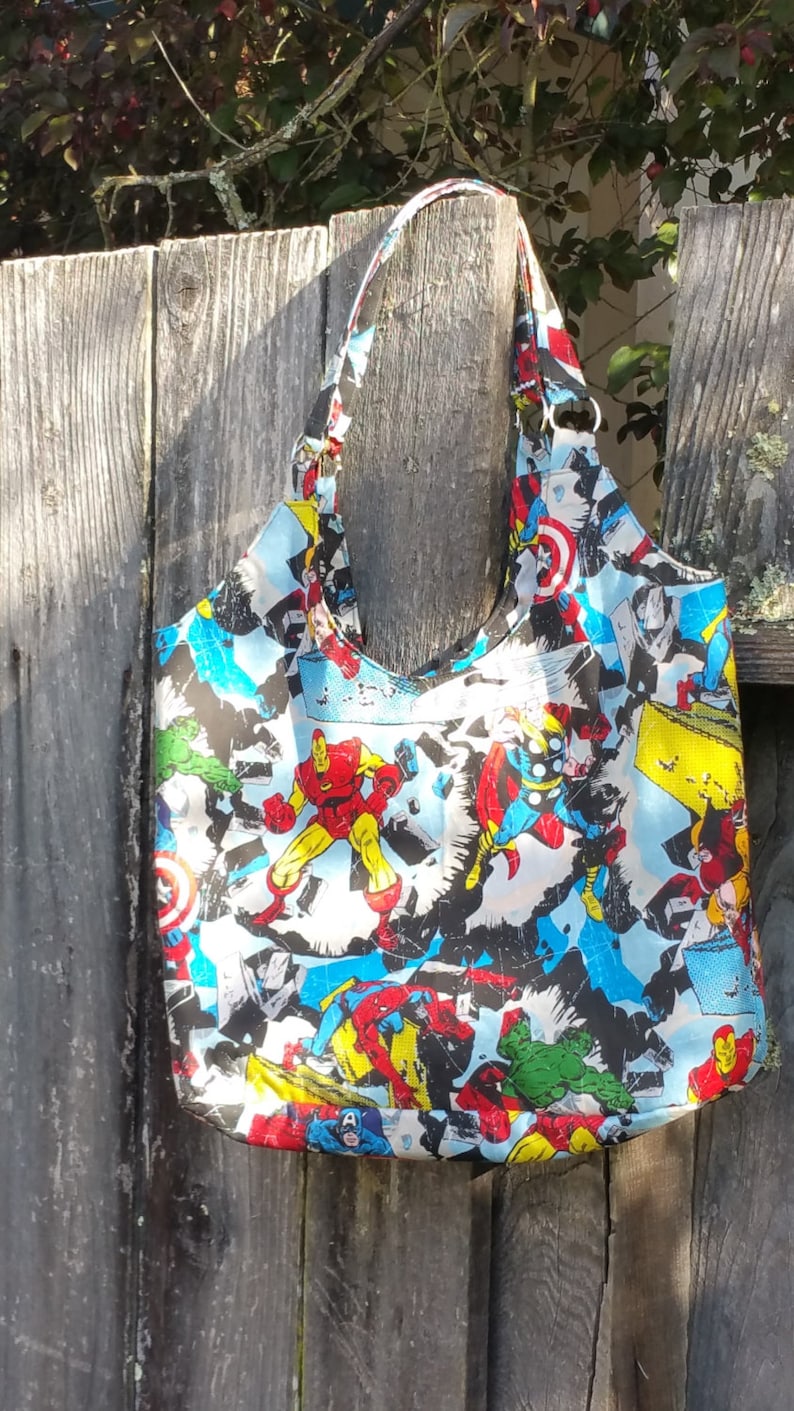 May include: A black and white tote bag with a colorful print of various superheroes, including Iron Man, Captain America, Thor, and Hulk. The bag has a long strap and is made of a durable fabric.
