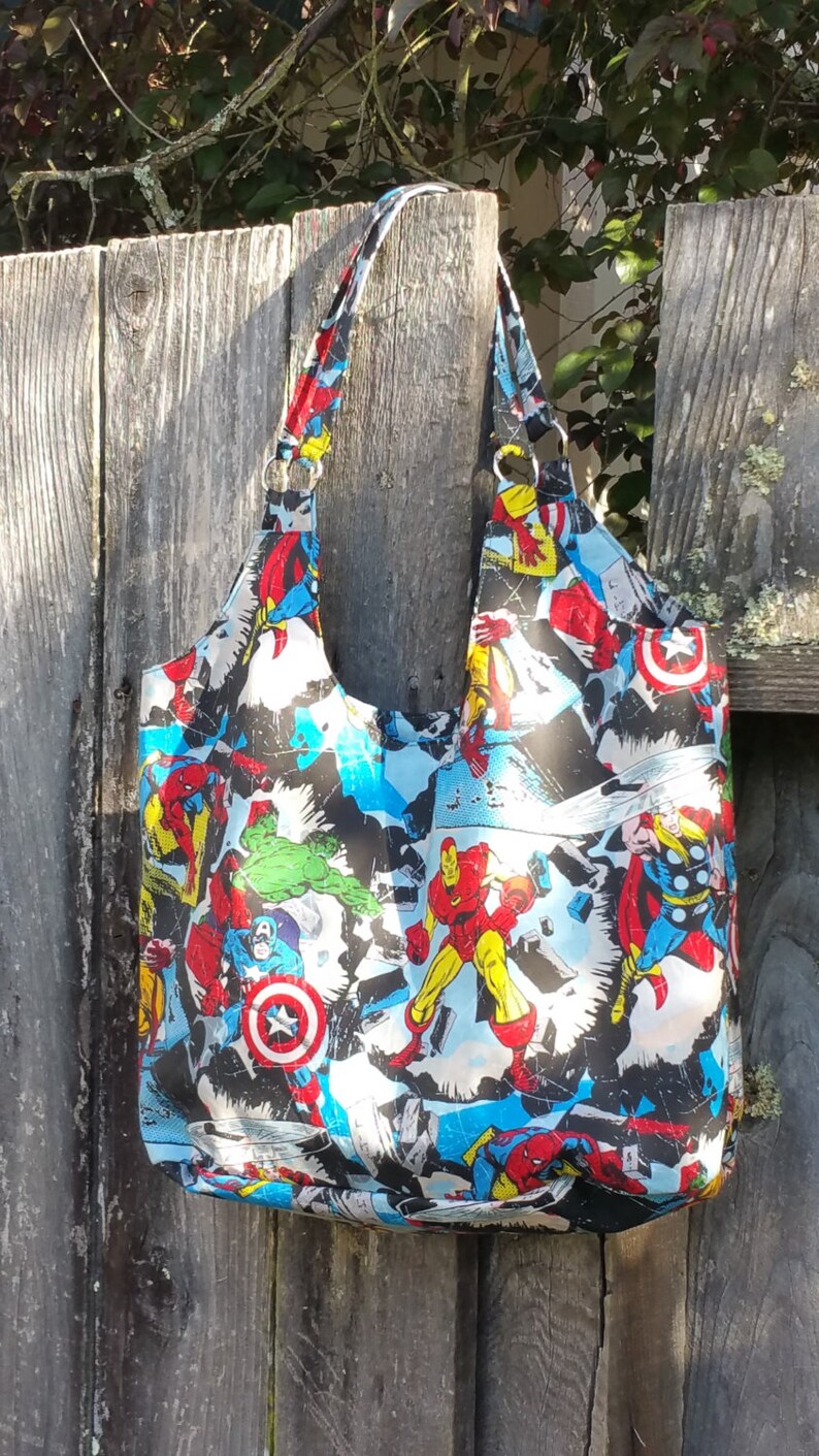 May include: A black tote bag with a colorful comic book print featuring superheroes like Iron Man, Captain America, and Thor. The bag has two long straps.