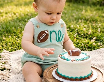 May include: A light sage green ribbed romper featuring two wooden buttons at the shoulders and an embroidered brown football with the word one in teal cursive script. The romper is paired with a white frosted cake topped with a small football.