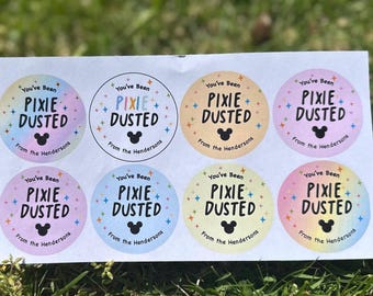 Puede incluir: Una hoja de ocho pegatinas circulares con el texto "You've Been Pixie Dusted" en una fuente caprichosa, con un gráfico de la cabeza de Mickey Mouse. Cada pegatina tiene un esquema de color pastel diferente y el texto "From the Hendersons".