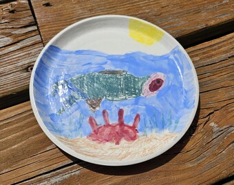 May include: A hand-painted ceramic plate featuring an underwater scene. The design includes a green sea turtle, a red crab, blue water, and a yellow sun. The plate has a white rim and is set on a wooden surface.