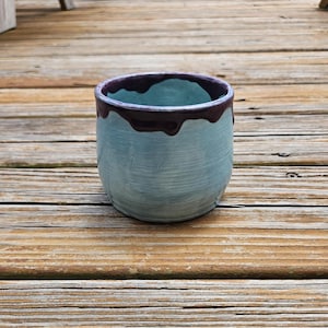 May include: A ceramic cup with a light blue, textured body and a dark brown, glossy glaze dripping from the rim down the sides. The vessel has a rounded shape and a smooth interior, showcasing a contrast between the matte blue and dark brown.