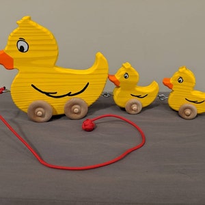 May include: A wooden pull-along toy featuring a large yellow duck leading two smaller ducklings. The ducks have orange beaks, black eyes, and are connected by a chain. A red cord with a knot is attached to the lead duck.