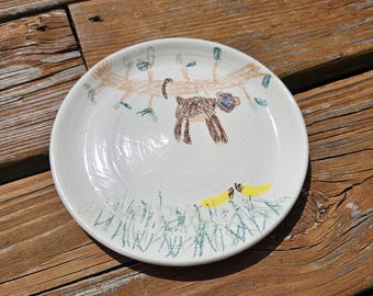 May include: White ceramic plate with a hand-drawn illustration of a brown animal under a tree with green leaves. The plate also features green grass and yellow objects. The artwork is in brown, blue, green, and yellow.
