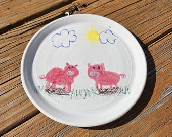 May include: White ceramic plate with a hand-drawn design featuring two pink pigs, blue clouds, and a yellow sun. The plate has a simple, whimsical style, perfect for serving food or as a decorative piece.