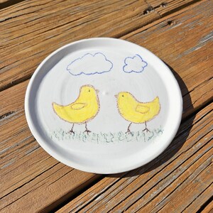 May include: A white ceramic plate with a hand-painted design of two yellow chicks standing on green grass with blue clouds in the background.