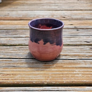 May include: A ceramic cup with a rounded, bulbous shape featuring a two-tone glaze. The upper portion is a deep, dark purple that transitions into a soft, muted pink on the lower half, with visible horizontal ridges along the body of the vessel.