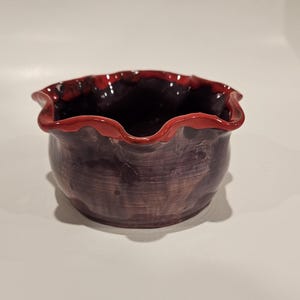 May include: A small, dark purple ceramic bowl with a red, wavy rim. The bowl has a rustic, handmade look.