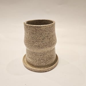 May include: A speckled ceramic vase with a cylindrical shape and a small, round base. The vase has a light beige color with dark brown speckles throughout. The design includes a slight indentation in the middle, adding a unique touch to the pottery.
