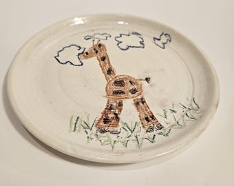 May include: A white ceramic plate with a hand-painted giraffe design. The giraffe has brown spots and is standing in green grass under blue cloud illustrations. The plate is round and has a slightly raised edge.